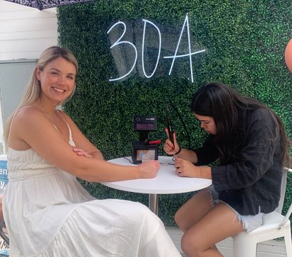 Smiling woman in a white sundress getting nail art from an artist at a small outdoor patio table under a patterned umbrella, with a green boxwood wall and neon "30A" sign behind them.