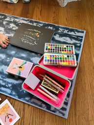 At-home art setup on a hardwood floor and patterned mat: open watercolor palettes, colorful pens and markers in a pink-lined case, a floral crescent-moon sketchbook, a pastel card box, and a bare foot with pink toenails.