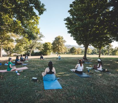 Outdoor yoga class in a riverside park on a sunny day, participants seated on mats spaced under large shade trees with river and rolling hills in the background.