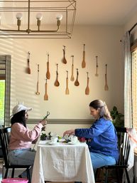 Two people arranging flowers at a small dining table in a cozy breakfast nook, with a wall display of hanging wooden spoons, a brass cage chandelier overhead, and sunlit window blinds.