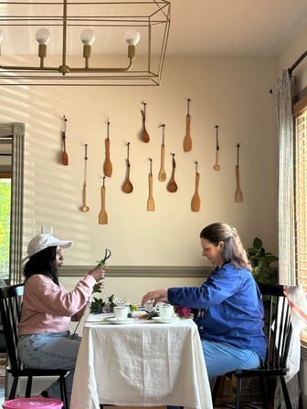 Two people arranging flowers at a small dining table in a cozy breakfast nook, with a wall display of hanging wooden spoons, a brass cage chandelier overhead, and sunlit window blinds.