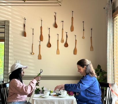 Two people arranging flowers at a small dining table in a cozy breakfast nook, with a wall display of hanging wooden spoons, a brass cage chandelier overhead, and sunlit window blinds.