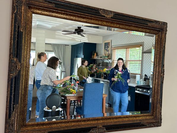 Ornate wall mirror reflecting a bright home kitchen where a small group arranges colorful flowers on a wooden dining table beside blue cabinets, patterned wallpaper and wooden blinds.