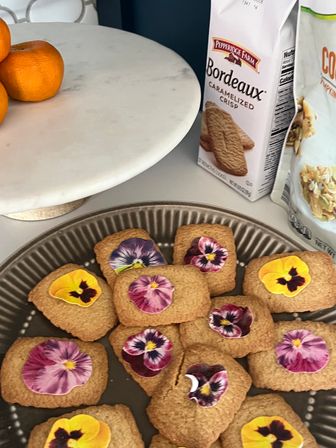 Tray of square shortbread cookies decorated with vibrant edible pansy flowers on a kitchen counter beside a marble cake stand with mandarins and a box of caramelized crisp cookies.
