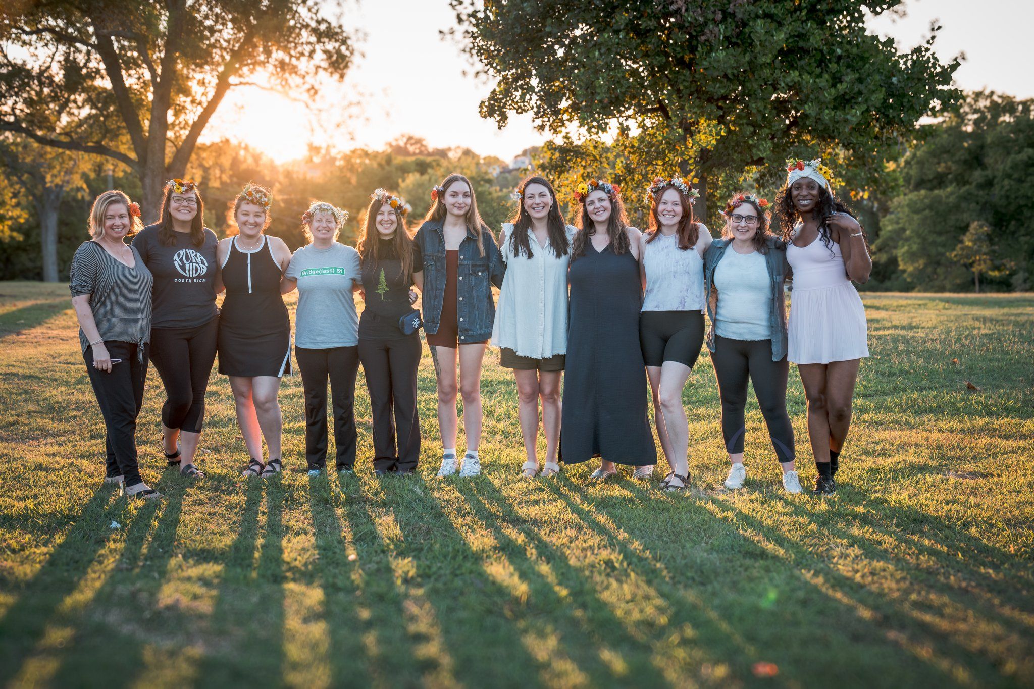 Group of twelve women wearing flower crowns stand arm-in-arm in a grassy park at sunset, smiling and backlit by warm golden-hour light.