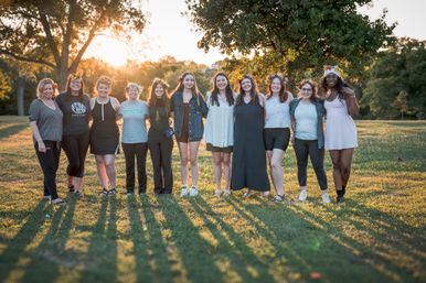 Group of twelve women wearing flower crowns stand arm-in-arm in a grassy park at sunset, smiling and backlit by warm golden-hour light.