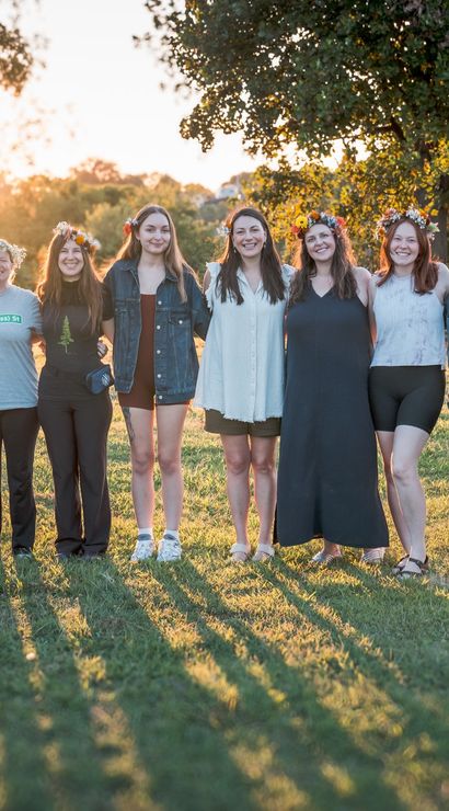 Group of twelve women wearing flower crowns stand arm-in-arm in a grassy park at sunset, smiling and backlit by warm golden-hour light.