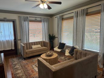 Cozy sunlit living room with beige velvet sofas, large wooden blinds and patterned rug, round wooden coffee table, accent chairs with fluffy throws and a ceiling fan
