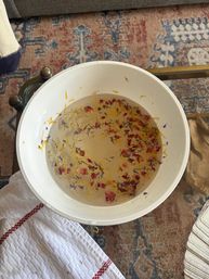 White bowl of water with floating dried flower petals in yellow, pink and blue sitting on a glass coffee table over a patterned rug in a cozy living room.