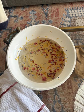 White bowl of water with floating dried flower petals in yellow, pink and blue sitting on a glass coffee table over a patterned rug in a cozy living room.