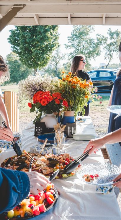 Sunny park picnic under a wooden pavilion with people serving from buffet platters, fresh fruit and appetizers, and a bright flower centerpiece — casual summer gathering.