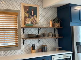 Modern kitchen corner with navy cabinets and white arabesque tile backsplash; wooden blinds, two rustic wood shelves holding stacked gray dishes, glasses, wood bowls, cookbooks, a copper pitcher and a potted plant, knife block and storage jars on a white quartz countertop, and a framed vintage portrait on the top shelf.