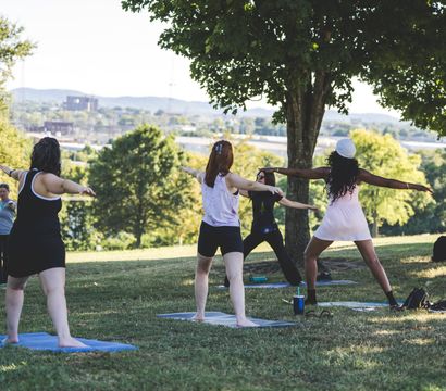 Outdoor yoga class in a sunlit park, students in warrior pose on mats beneath trees with a city skyline and rolling hills in the background.