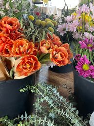 Vibrant flower shop display: orange ranunculus bouquets, yellow billy balls, pink daisies and purple mums in black buckets with eucalyptus sprigs on a wooden floor
