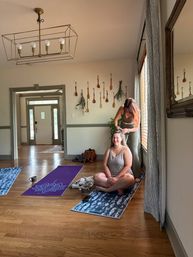 Two women in a bright home yoga room with hardwood floors and colorful mats — one sits cross-legged smiling while the other braids her hair near a sunlit window, decorative wooden spoons on the wall and a modern brass chandelier overhead.