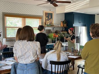 Sunlit home kitchen workshop — instructor leading a small flower-arranging class by navy cabinets, patterned tile backsplash and wooden blinds.