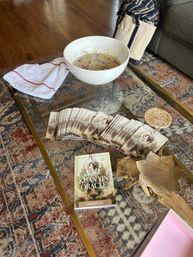 Cozy living room scene on a patterned rug: glass coffee table holding a Kuanyin Oracle guidebook and fanned oracle cards, a white bowl with floating flower petals, and decorative wooden and brass accents.
