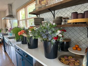 Sunlit kitchen counter with blue cabinets and white arabesque tile backsplash, open wooden shelves of dishes, black buckets of fresh mixed flowers on the white countertop, a plate of floral-decorated cookies and a small stack of tangerines.