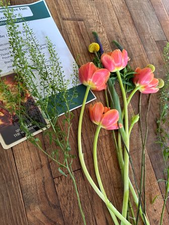 Coral-pink tulips, yellow billy balls and green foliage laid on a rustic wood tabletop next to a floral workshop flyer