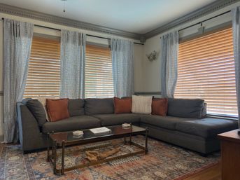 Cozy sunlit living room corner with a gray L-shaped sectional, rust and cream throw pillows, wooden horizontal blinds, patterned sheer curtains, glass coffee table and a vintage-style area rug.