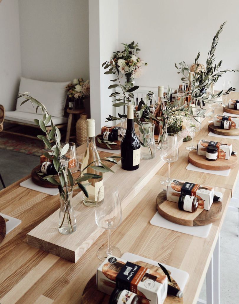 Sunlit modern-rustic dining table set for wine tasting or brunch with bottles, empty wine glasses, olive-branch vases and wooden boards of snacks.