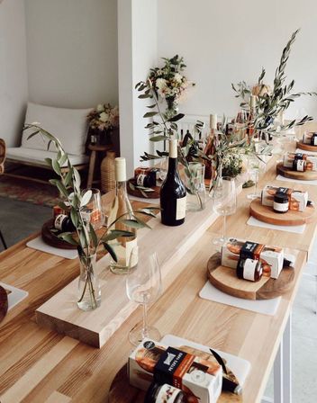 Sunlit modern-rustic dining table set for wine tasting or brunch with bottles, empty wine glasses, olive-branch vases and wooden boards of snacks.