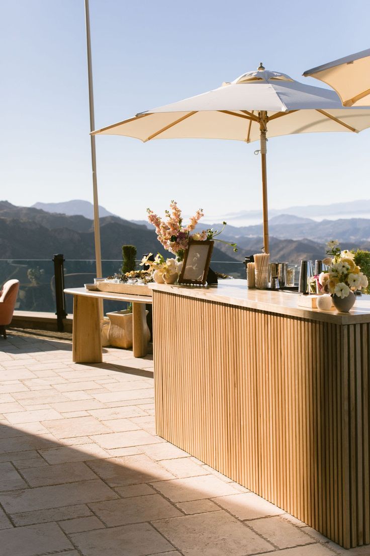 Al fresco wooden bar on a sunlit mountain terrace with cream patio umbrellas, fresh floral centerpieces and cocktail tools overlooking layered mountain ranges under a clear blue sky