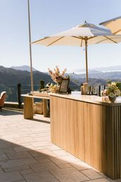 Al fresco wooden bar on a sunlit mountain terrace with cream patio umbrellas, fresh floral centerpieces and cocktail tools overlooking layered mountain ranges under a clear blue sky