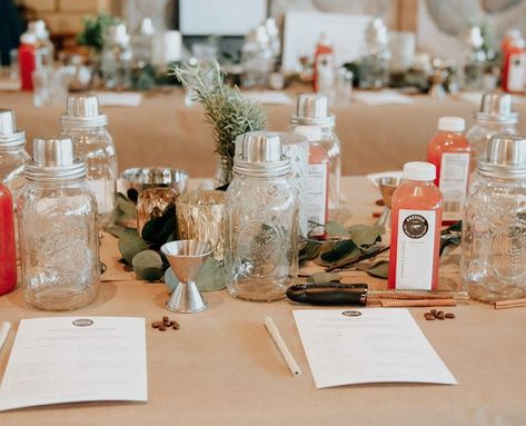 Farmhouse-style workshop table set for a DIY cocktail-mixing class with mason jars, jiggers, bottled mixers, recipe cards and a greenery centerpiece.