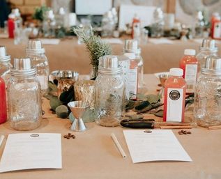 Farmhouse-style workshop table set for a DIY cocktail-mixing class with mason jars, jiggers, bottled mixers, recipe cards and a greenery centerpiece.