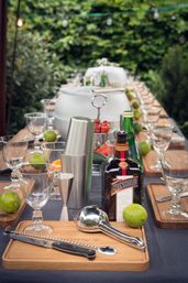 Outdoor garden cocktail station with wooden boards, glassware, fresh limes, stainless steel shaker and orange liqueur bottle, mixology tools neatly arranged
