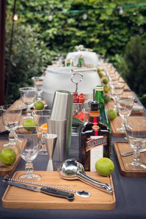Outdoor garden cocktail station with wooden boards, glassware, fresh limes, stainless steel shaker and orange liqueur bottle, mixology tools neatly arranged
