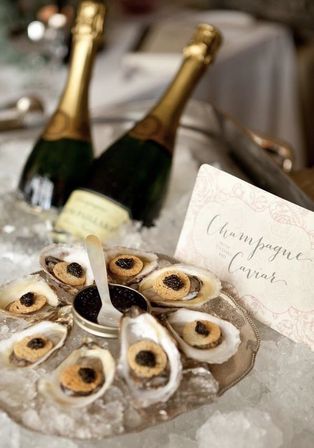 Oysters on the half shell topped with caviar and small toasted rounds on crushed ice, an open tin of black caviar and two chilled champagne bottles behind a decorative “Champagne & Caviar” card.
