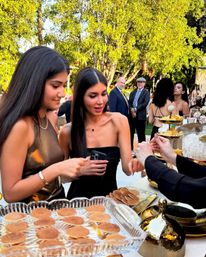 Two women in elegant black dresses sample mini pancakes at an outdoor garden reception dessert table, guests mingling under sunlit trees in the background