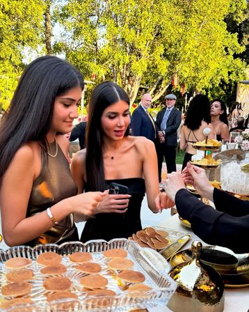 Two women in elegant black dresses sample mini pancakes at an outdoor garden reception dessert table, guests mingling under sunlit trees in the background