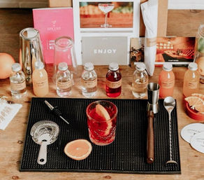 Mixology station on a wooden bar: bright red cocktail with a grapefruit slice on a black bar mat, surrounded by jigger, bar spoon, Hawthorne strainer, pourer and small labeled bottles of juices and spirits.