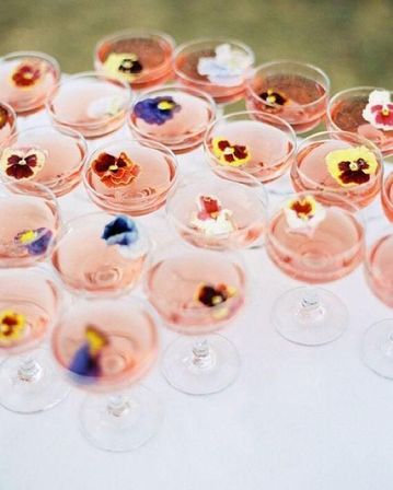 Pink cocktails in coupe glasses topped with colorful edible pansy flowers, arranged as a festive wedding or garden party drink display