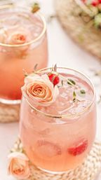 Close-up of a pink floral cocktail in a gold-rimmed glass filled with ice, raspberries and garnished with peach roses on a woven coaster — refreshing garden-party drink.