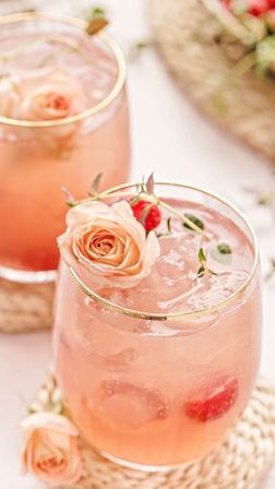 Close-up of a pink floral cocktail in a gold-rimmed glass filled with ice, raspberries and garnished with peach roses on a woven coaster — refreshing garden-party drink.