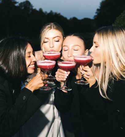Four women toasting with pink frothy cocktails in coupe glasses at an outdoor evening gathering