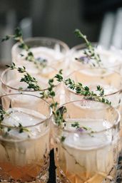 Six crystal lowball cocktails with large square ice cubes and fresh thyme sprigs, pale peach craft drinks styled for a summer brunch or celebration.
