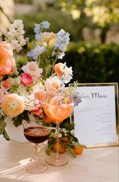 Sunlit garden wedding bar setup with pastel floral centerpiece, a bright citrus spritz in a goblet with striped straw, a dark espresso-style cocktail in a coupe, framed bar menu and fresh tangerines on a wooden table