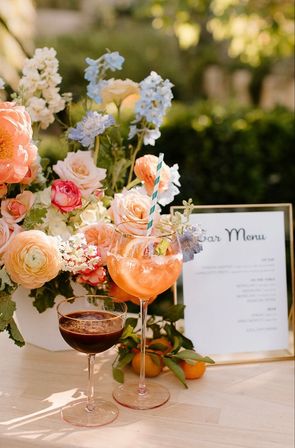 Sunlit garden wedding bar setup with pastel floral centerpiece, a bright citrus spritz in a goblet with striped straw, a dark espresso-style cocktail in a coupe, framed bar menu and fresh tangerines on a wooden table