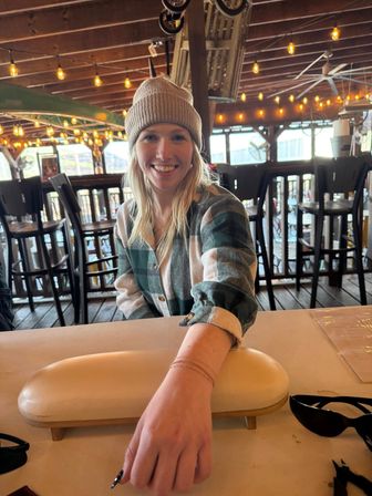 Smiling woman in a knit beanie and green plaid shirt reaching across a table at a cozy rustic covered patio bar with warm string lights, wooden beams, and high stools in the background.