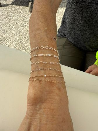 Sun-spotted forearm resting on a light cushion wearing a stack of delicate gold and silver chain bracelets, photographed outdoors on a textured patio with a person in a gray shirt in the background