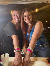 Two smiling women leaning on a vendor counter at an outdoor night market tent, lit by warm hanging bulbs, wearing colorful wristbands, bracelets and casual summer outfits.