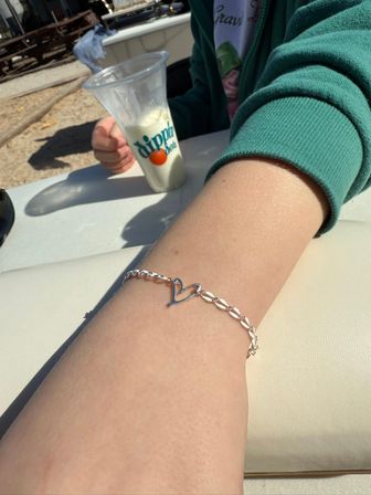 Sunlit close-up of a wrist wearing a delicate silver heart bracelet, person in a green sweatshirt holding a clear plastic cup of white frozen dessert at an outdoor patio seating area.
