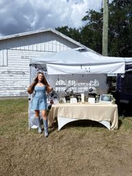 Smiling vendor in denim dress and silver cowboy boots at an outdoor craft market booth under a white canopy, table of handmade jewelry and decor set up in front of a weathered white barn.