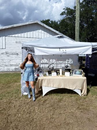Smiling vendor in denim dress and silver cowboy boots at an outdoor craft market booth under a white canopy, table of handmade jewelry and decor set up in front of a weathered white barn.