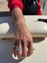 Sunlit close-up of a hand resting on a white surface outdoors, wearing stacked diamond rings, a delicate bracelet and blue-painted nails with a red sleeve in the background.